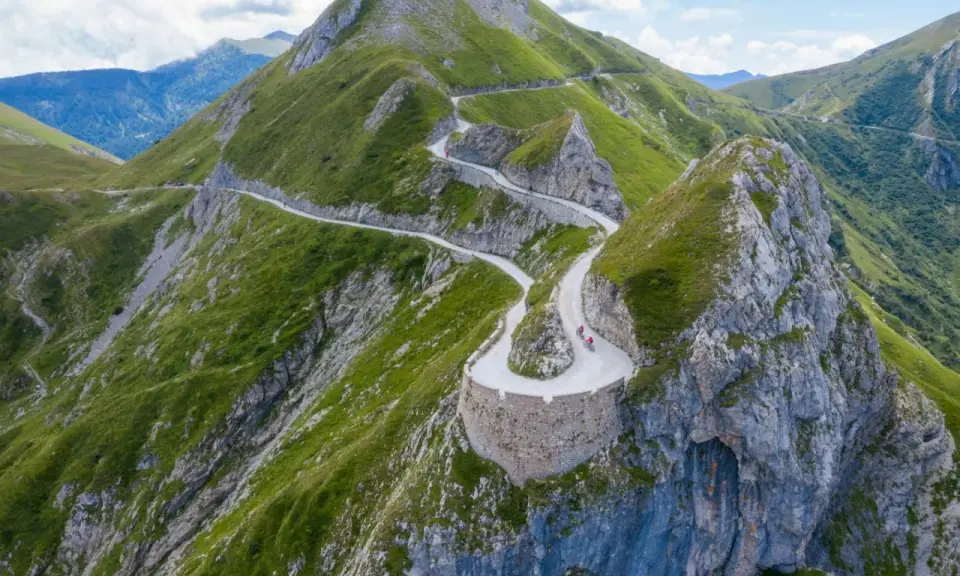 Un tortuoso cammino del sale si snoda tra montagne verdi, con ciclisti che affrontano tornanti mozzafiato su un sentiero panoramico.