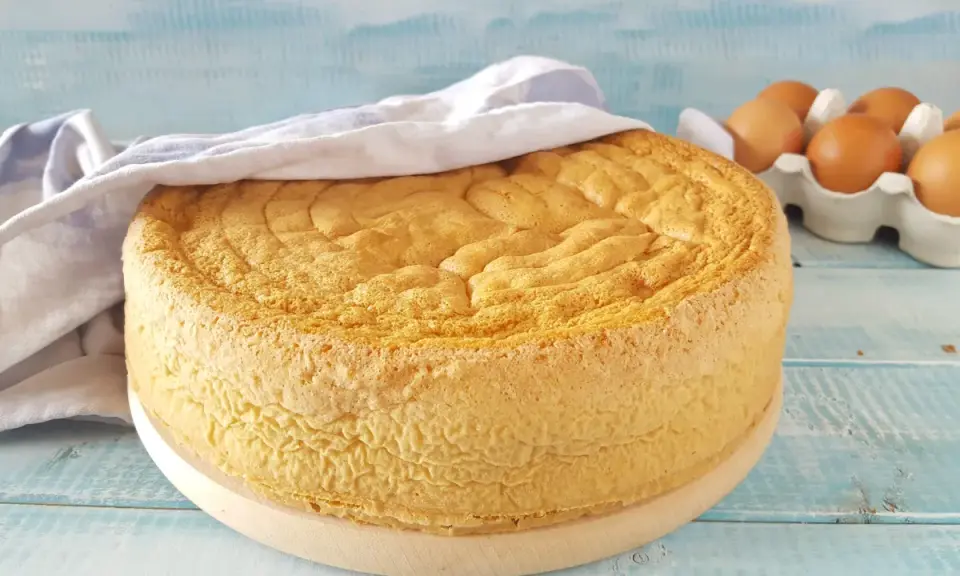 Un soffice pan di spagna dorato, coperto da un canovaccio, con uova fresche sullo sfondo.