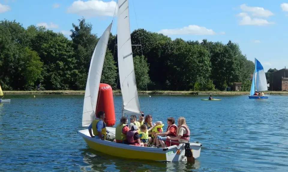 Un groupe de personnes, dont des enfants, profite d'une journée ensoleillée sur un bouchain bateau jaune. Un autre bateau à voile bleu navigue au loin.