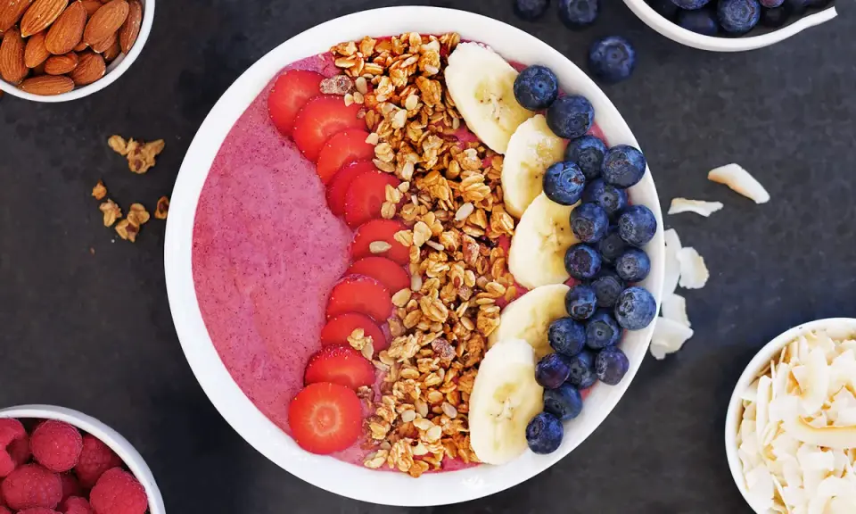 En färgglad acai bowl med jordgubbar, banan, blåbär och granola. Serverad med mandlar och kokosflingor vid sidan.