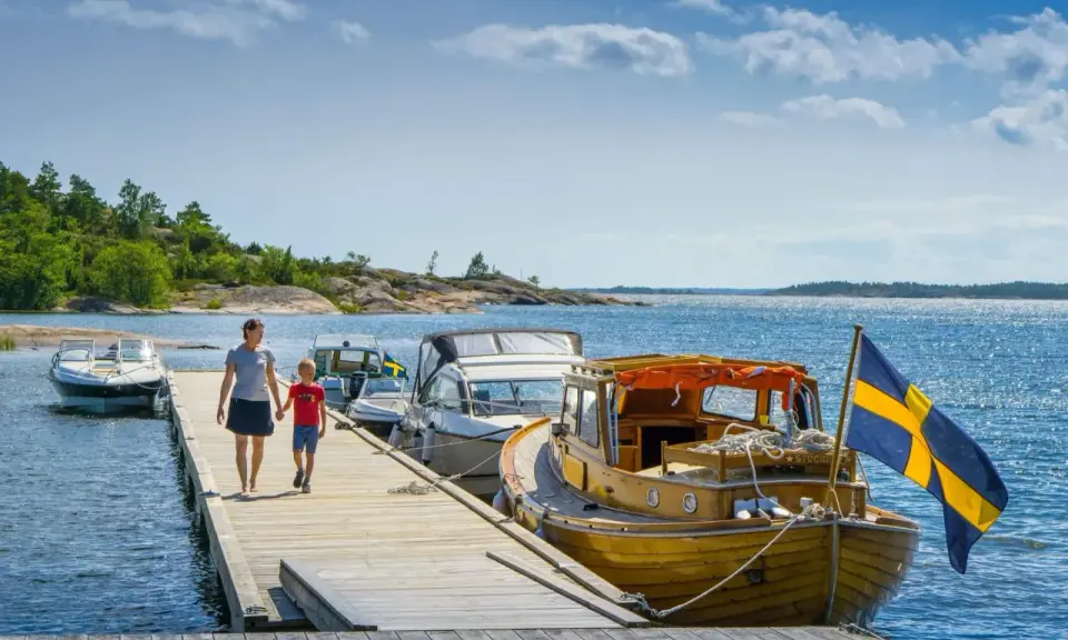 En kvinna och ett barn går på en brygga med båtar och en svensk flagga. En perfekt bild av en semester i Sverige.
