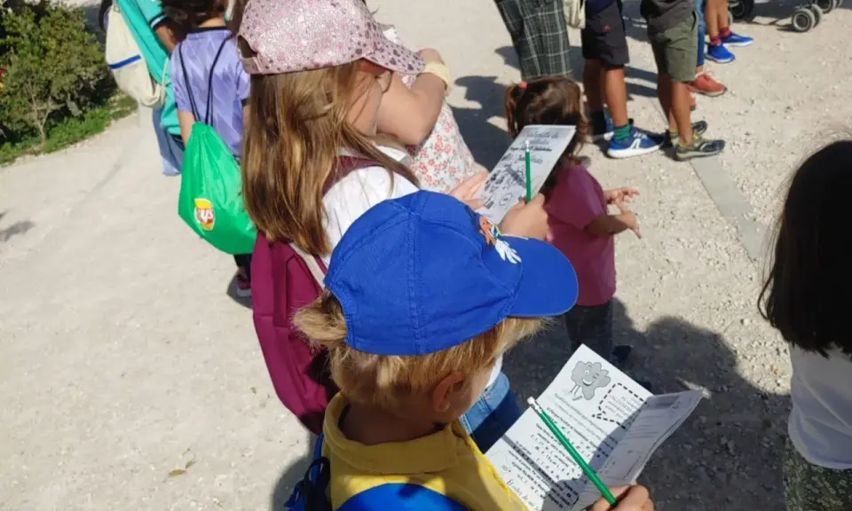 Niños con gorras y mochilas, algunos con folletos, explorando el **parque de Valdebebas** en un día soleado.