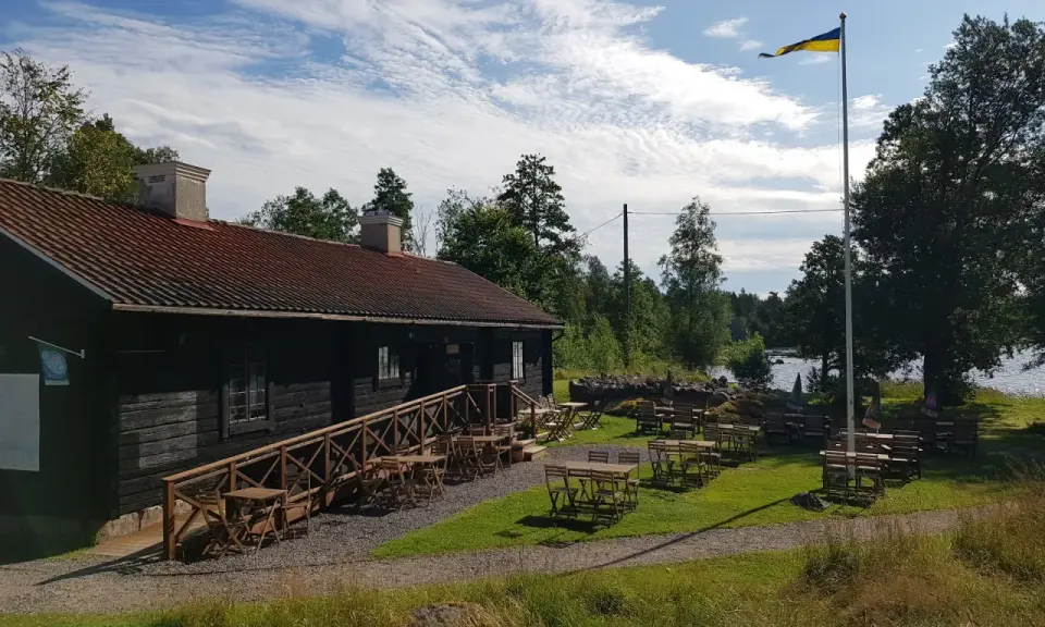 Mysigt tiveden boende med uteservering vid vattnet. En svensk flagga vajar i vinden.