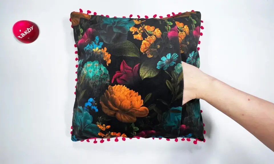 Ett sammetslent kuddfodral med en mörk blommig design och en lekfull kant av små röda bollar. En hand sticker fram ur en ficka på kudden.