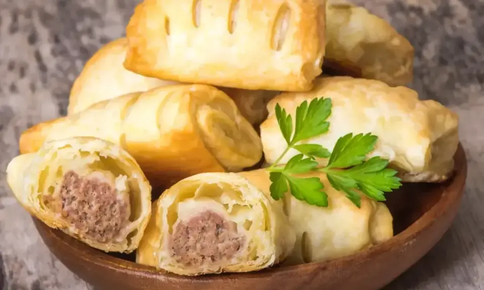 Stos pieczonego ciasta francuskiego z kiełbaską w środku, ozdobiony gałązką pietruszki.