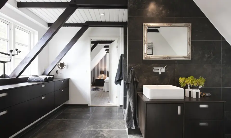 Baño moderno con vigas oscuras y suelo gris. El lavabo sobre encimera y los muebles oscuros contrastan con las paredes de baldosas oscuras. Se ve un dormitorio al fondo.
