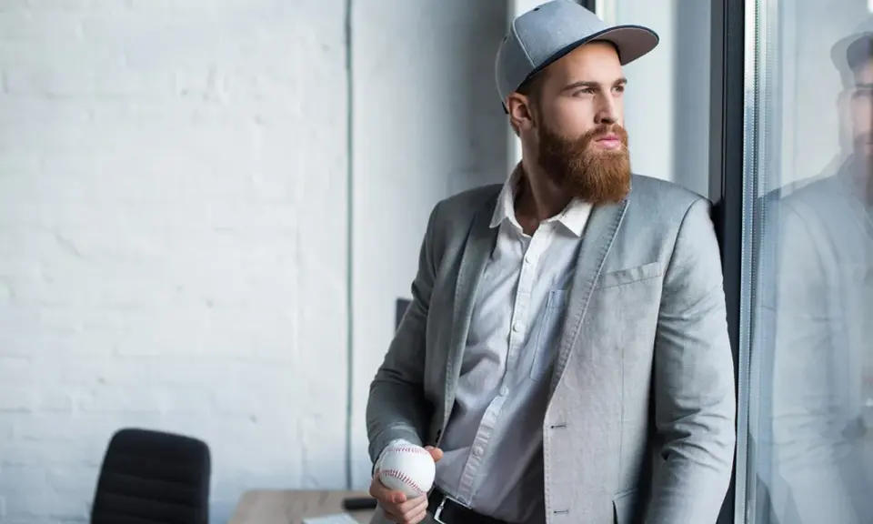 Mężczyzna w szarej czapce snapback i garniturze trzyma piłkę baseballową.