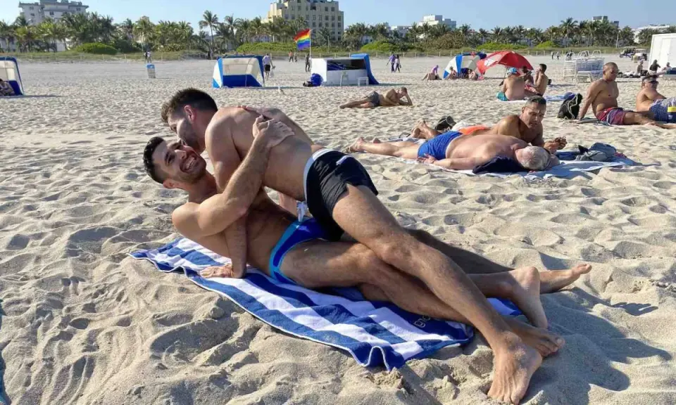 Two men embrace on a towel at Haulover Beach, a popular spot for gay beachgoers. Other sunbathers relax on the sand in the background.