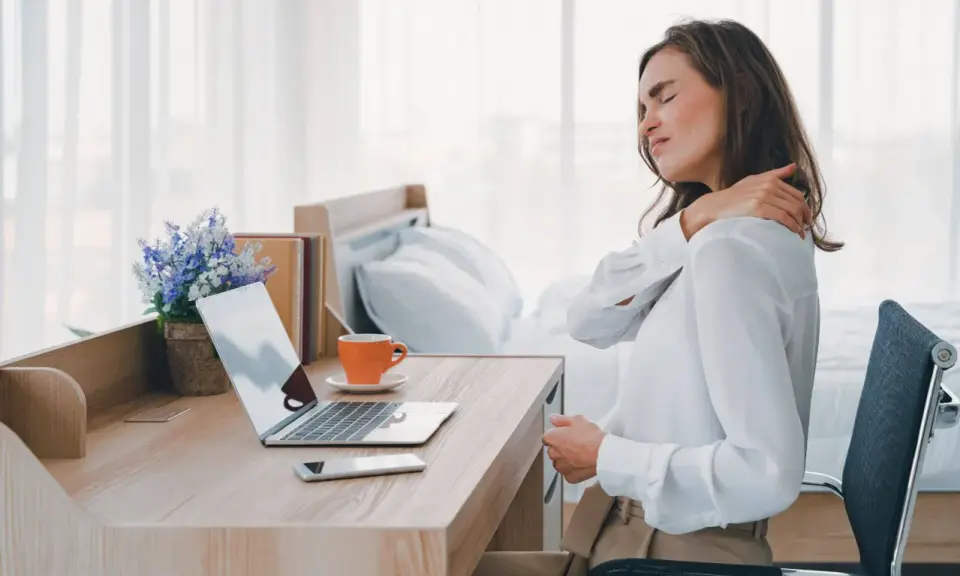 Femme se massant l'épaule, assise à son bureau. La mauvaise position bureau peut causer des douleurs.