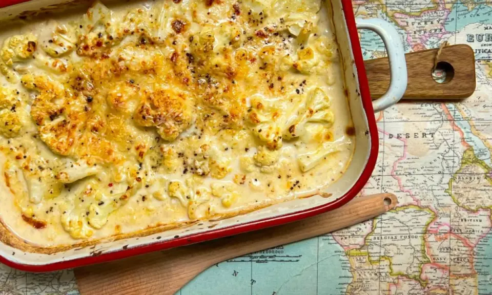 Coliflor gratinado con bechamel cremosa, cubierto de queso dorado y pimienta negra, servido en una fuente roja sobre un mapa antiguo.