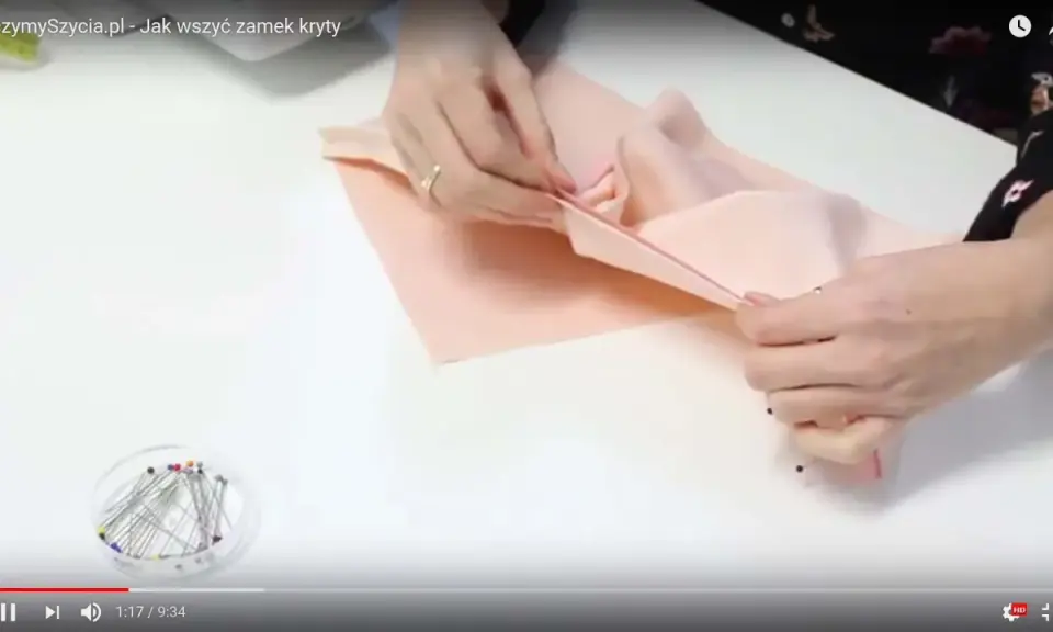 Ręce przygotowują materiał do szycia, pokazując, jak wszyć zamek kryty. Szpilki czekają w pojemniczku.