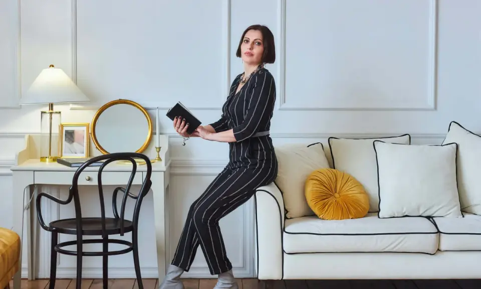 Elegancka kobieta w paski, siedząca na sofie, czyta książkę. Wnętrze z lampą i lustrem to kwintesencja stylu glamour.