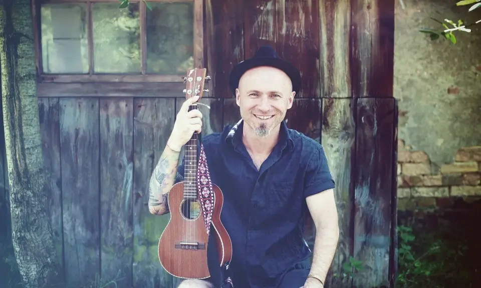 Uśmiechnięty mężczyzna w kapeluszu trzyma ukulele, gotowy do grania nim zajdzie słońce.