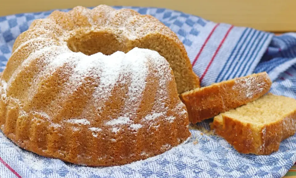 Puszysta babka wielkanocna, oprószona cukrem pudrem, leży na niebiesko-białym ręczniku. Obok niej kilka kromek ciasta.