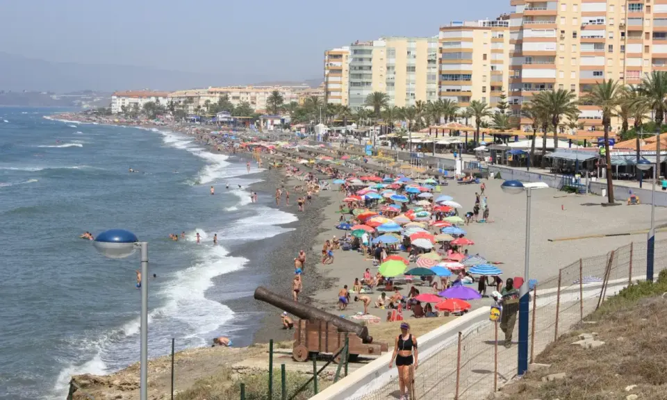 Sonniger Torrox Strand mit vielen bunten Sonnenschirmen, Menschen im Meer und an Land, sowie historischen Kanonen.