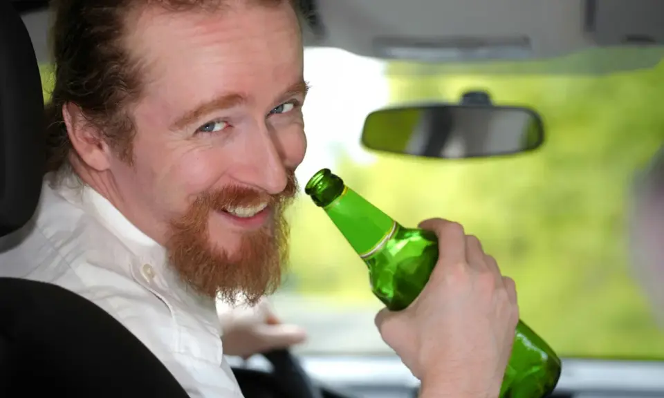 Uśmiechnięty mężczyzna z rudą brodą trzyma zieloną butelkę piwa w samochodzie. Mówi, że piwo to nie alkohol, ale jazda z nim jest niebezpieczna.