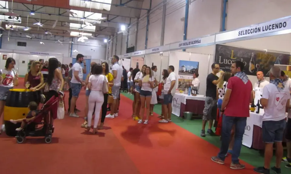 Gente en un evento de degustación de vinos, con stands de bodegas como "Coop Vinícola de Valdepeñas". El "túnel del vino Valdepeñas" se siente en el ambiente.