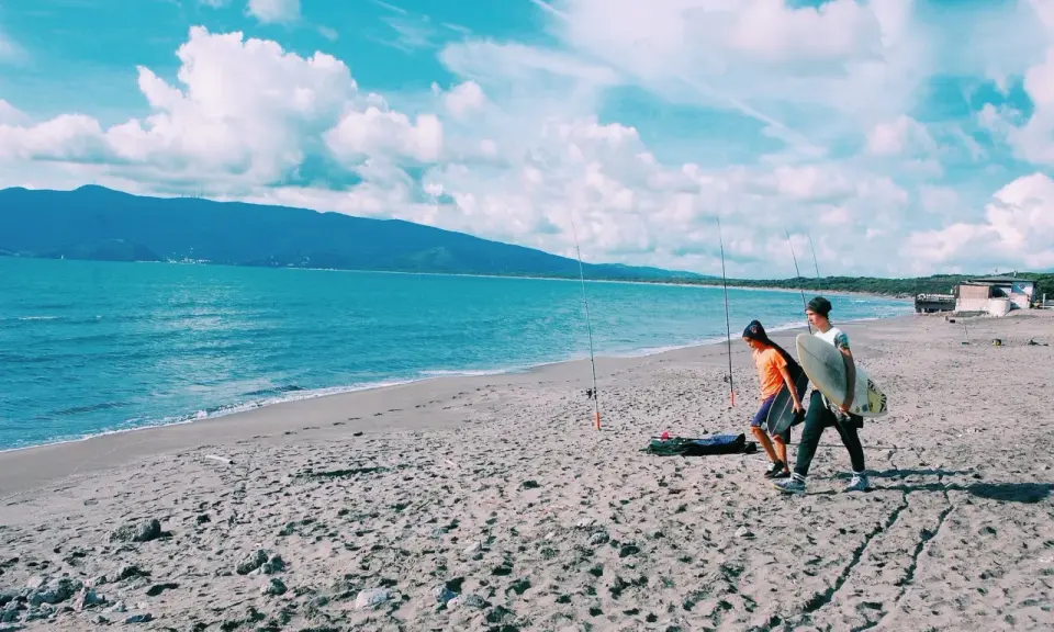 Due ragazzi passeggiano sulla spiaggia della Maremma Toscana, uno con una tavola da surf, mentre il mare azzurro lambisce la riva.