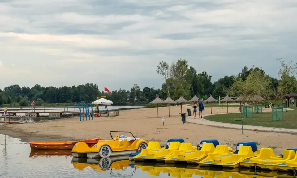 Żółte rowerki wodne i łódki czekają na plaży nad jeziorem blisko Warszawy. Para spaceruje po piasku pod słomianymi parasolami.