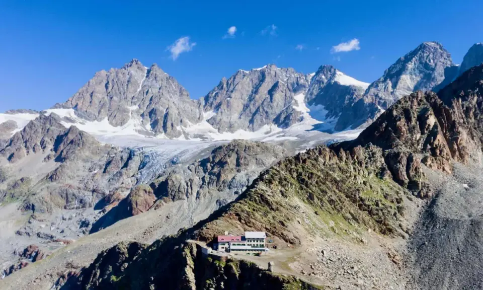 Il rifugio Marinelli, un edificio bianco e rosso, si erge su una cresta rocciosa con imponenti montagne innevate sullo sfondo.