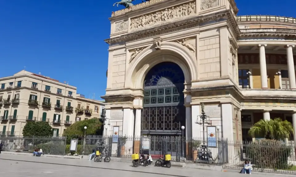 Il **Teatro Politeama Garibaldi** di Palermo, con la sua imponente facciata neoclassica, si staglia sotto un cielo azzurro. Tre scooter elettrici gialli sono parcheggiati davanti.