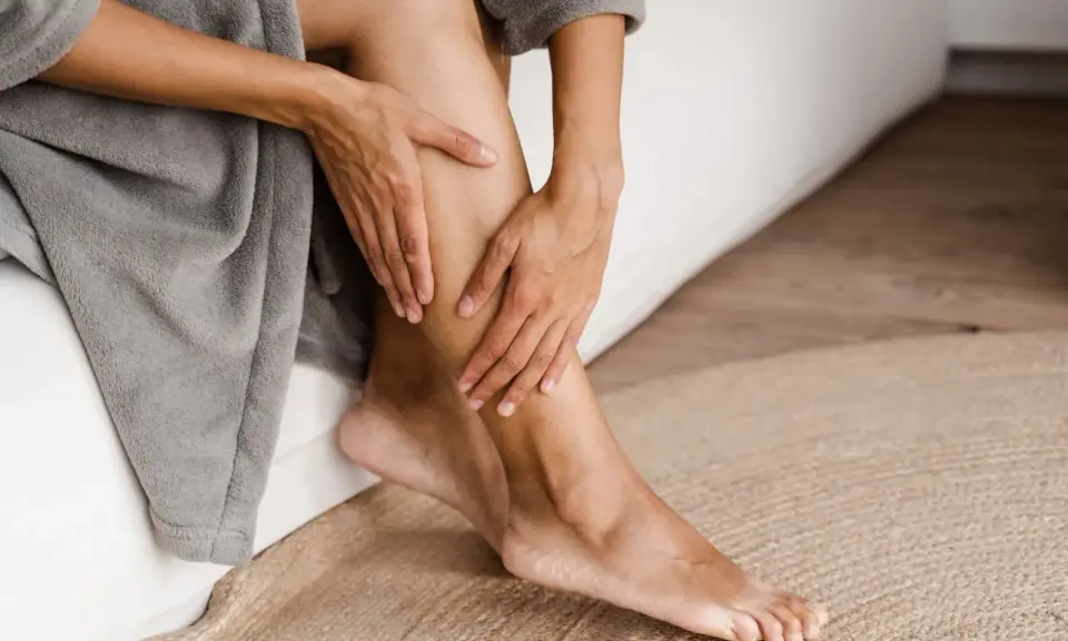 Mani che accarezzano le gambe, evidenziando la pelle secca. La persona è seduta su un letto, avvolta in un accappatoio grigio.