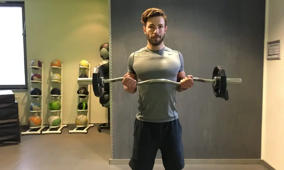 Mann macht Bizeps-Curls mit einer Langhantel. Im Hintergrund stehen Medizinbälle.