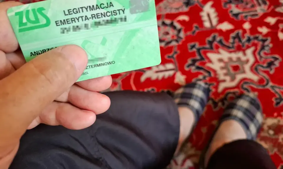 Zielona legitymacja ZUS "LEGITYMACJA EMERYTA-RENCISTY" w dłoni. Na dywanie widać stopy w kapciach.