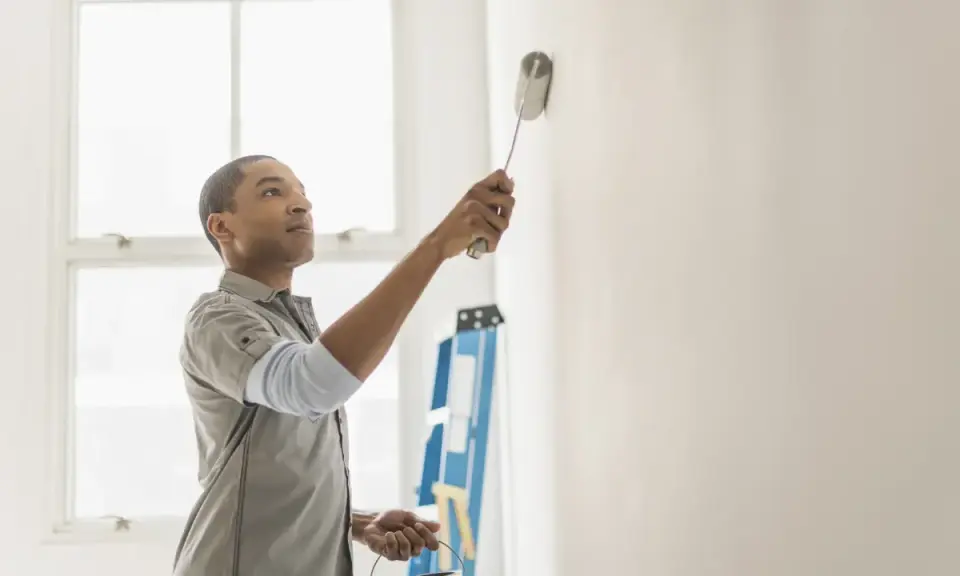 Hombre pintando pared blanca, resolviendo quién paga la pintura en un piso de alquiler. Escalera azul al fondo.