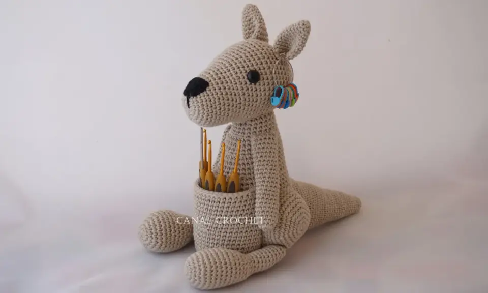 Canguro amigurumi beige sentado, con ganchillos en su bolsa y marcadores de puntos en su oreja.
