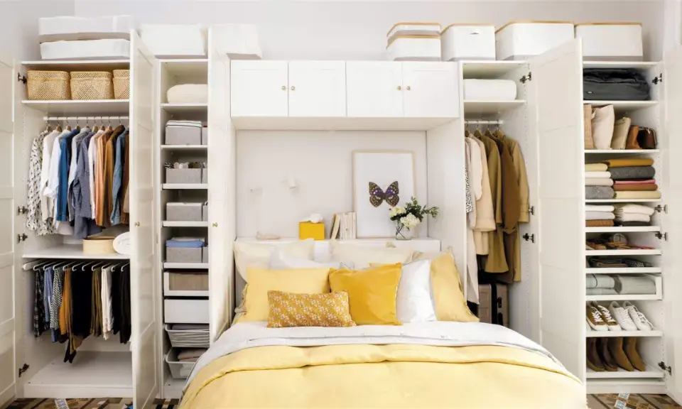 Habitación matrimonio pequeña con armario blanco, cama con ropa de cama amarilla y almohadas, y estanterías llenas de ropa y cestas.