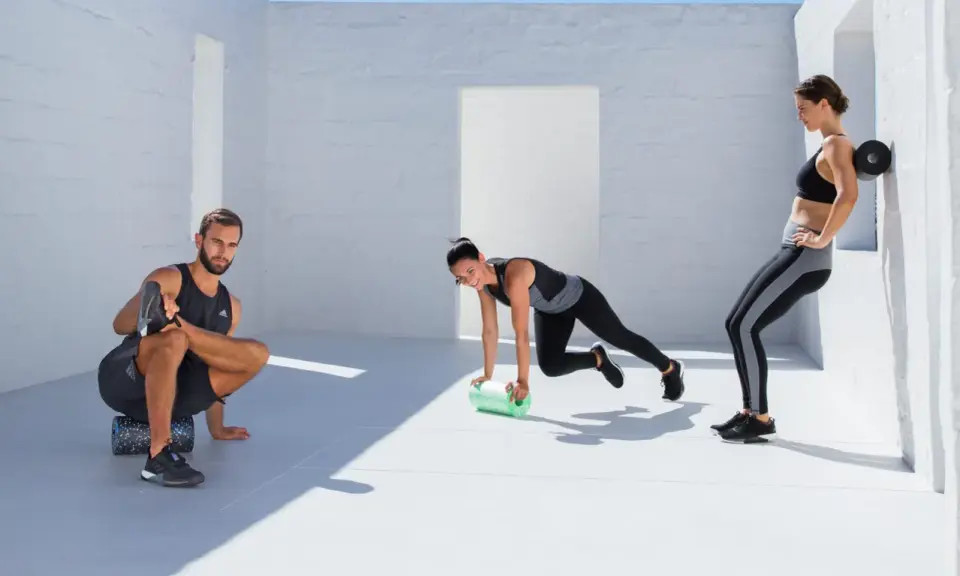 Drei Sportler nutzen eine **blackroll vor oder nach dem Training**. Ein Mann dehnt sich, eine Frau macht Mountain Climbers, die andere lehnt sich an die Wand.