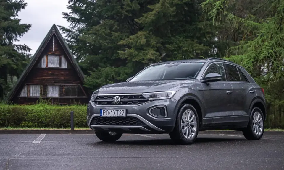 Szary Volkswagen T-Roc używany, zaparkowany przed drewnianym domkiem w lesie. Idealny na wypady za miasto.