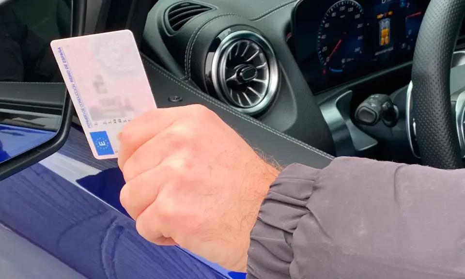 Mano sujetando un carnet de conducir español, con el interior de un coche moderno al fondo. Averigua el carnet b+e precio.