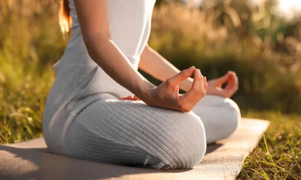 En person sitter i lotusställning på en matta utomhus, med händerna i meditation. Solen lyser genom gräset och skapar en varm atmosfär för morgonmeditation.