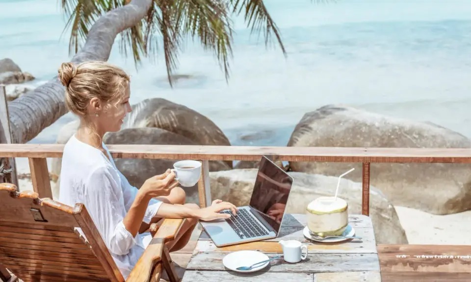 Kvinna med kaffe och laptop på en strandterrass, drömmen om ett digitalt nomad jobb.