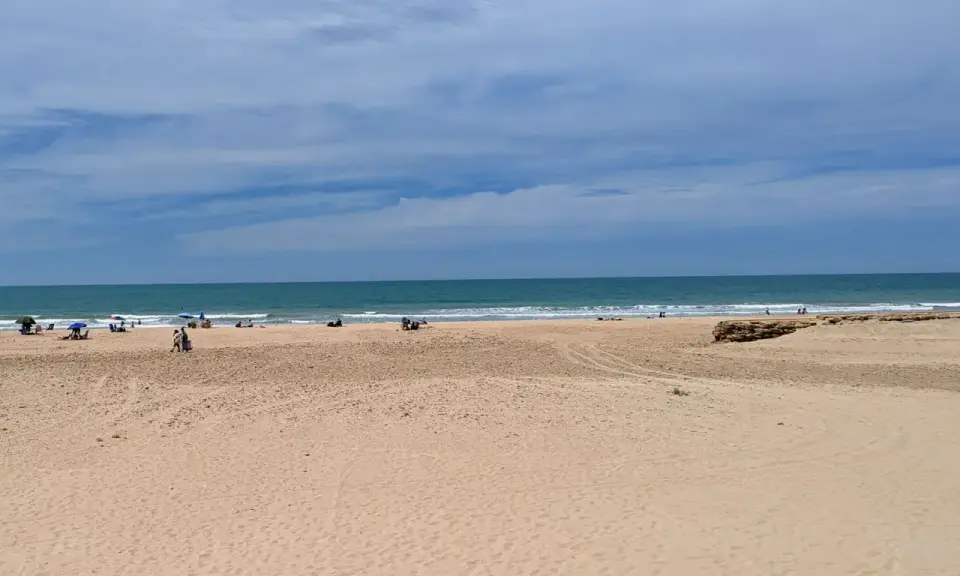 Weite Sandstrände und das Meer am Playa de Cortadura. Einige Menschen genießen den Tag unter Sonnenschirmen.