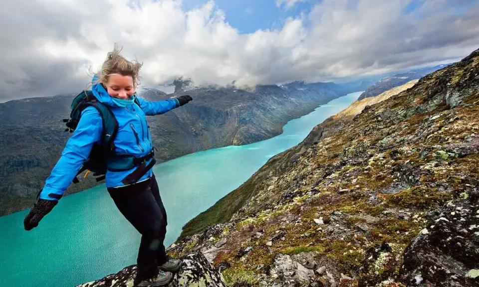 En kvinna med ryggsäck balanserar på en klippkant med en turkos sjö och berg i bakgrunden, en vy som påminner om Besseggen i Norge.