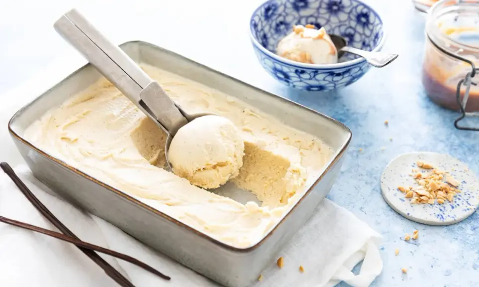Une cuillère prélève une boule d'une glace vanille maison crémeuse dans un plat. Des gousses de vanille et des éclats de cacahuètes sont à proximité.