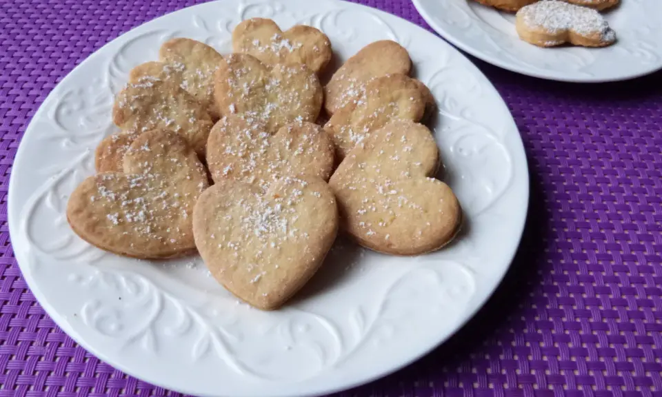 Stos ciasteczek cytrynowych w kształcie serc, posypanych cukrem pudrem, na białym talerzu.