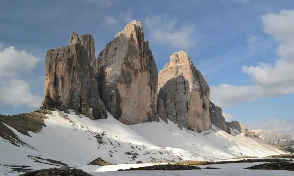 Majestatyczne **Tre Cime** w Dolomitach, ośnieżone zbocza i błękitne niebo z chmurami.