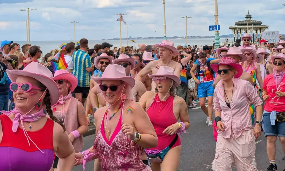 A vibrant Brighton gay pride parade, with many participants wearing pink cowboy hats and rainbow-themed attire, celebrating joyfully by the sea.
