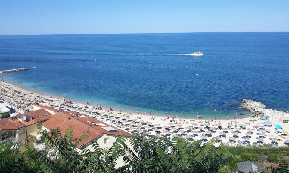 Le spiagge di Numana, con ombrelloni colorati e gente che si gode il sole, bagnate da un mare azzurro e calmo. Una barca solca l'orizzonte.