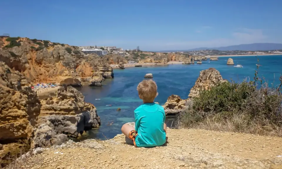 Kleinkind blickt auf die Küste Portugals. Felsen, türkisfarbenes Meer und eine Bucht mit Strand.