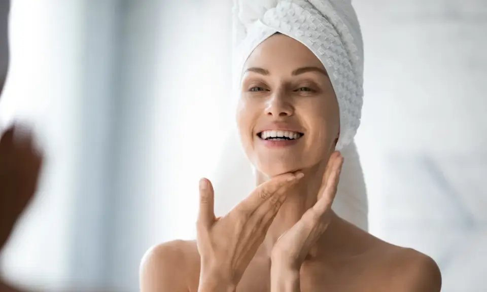 Une femme souriante, serviette sur la tête, applique délicatement de l'eau micellaire sur son visage pour une peau fraîche et nette.
