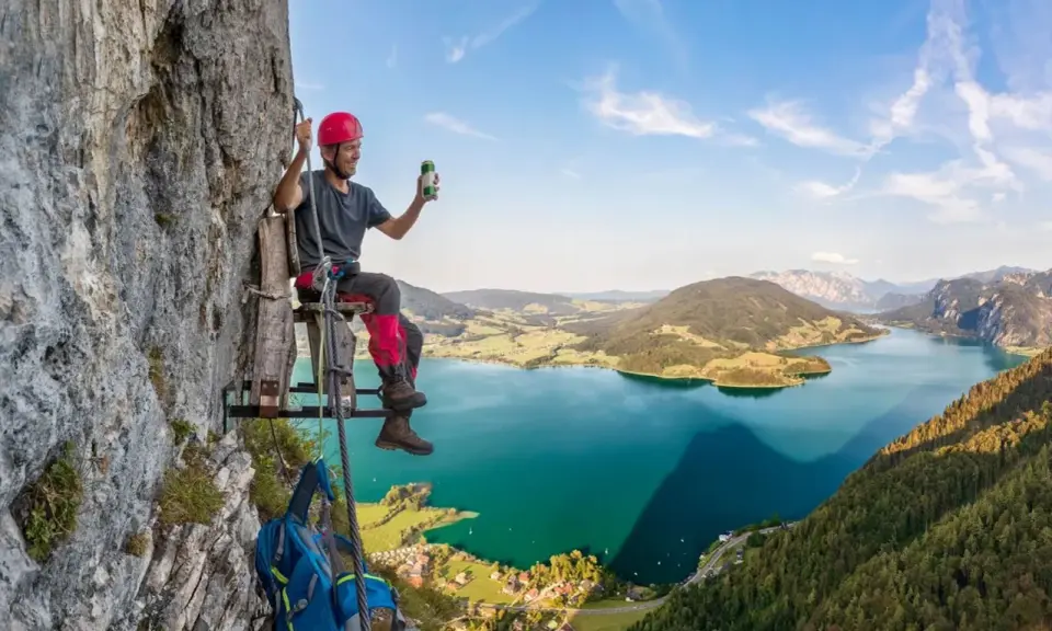 Mężczyzna w kasku siedzi na drewnianej platformie przy skalnej ścianie, ciesząc się widokiem na jezioro w Austrii. To wspaniała przygoda na via ferrata Austria!