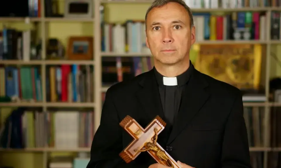 Duchowny z krzyżem w dłoniach, na tle regałów z książkami. Czy religia wlicza się do czerwonego paska?