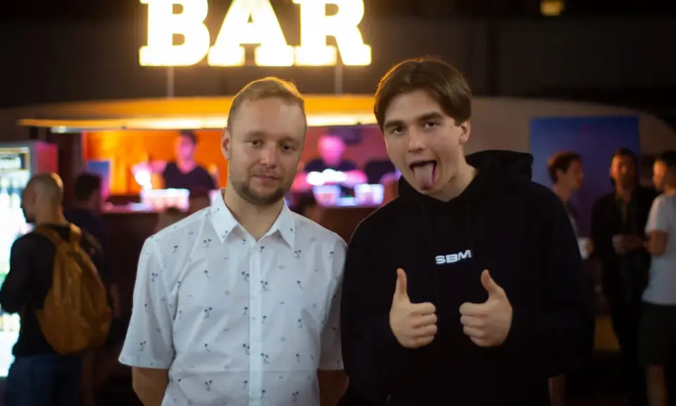 Dwóch mężczyzn pozuje przed napisem "BAR". Jeden w koszuli w palmy, drugi w bluzie z napisem SBM, pokazuje kciuki w górę i wystawia język.