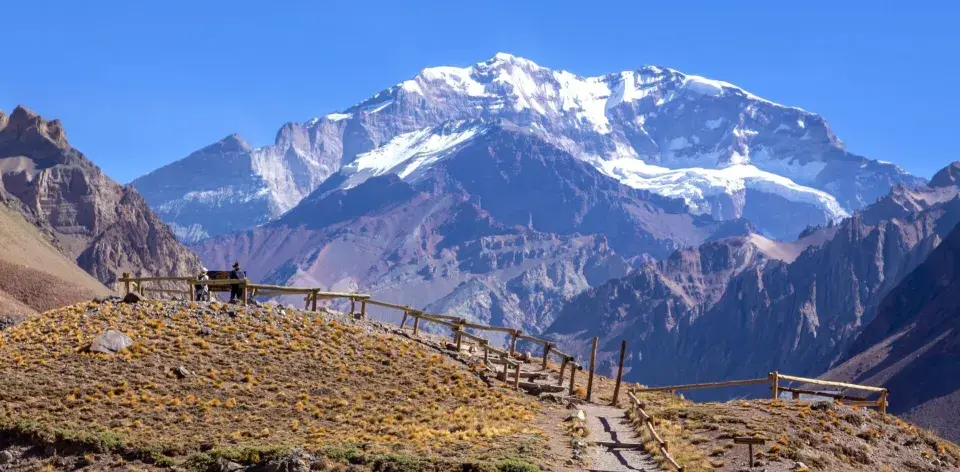 Widok na majestatyczne góry z ośnieżonym szczytem Aconcagua. Ścieżka z drewnianym ogrodzeniem prowadzi przez suchą, trawiastą przestrzeń.