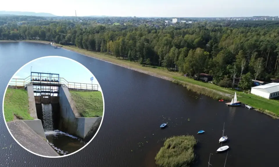 Widok na zbiornik wodny z zaporą i spływającą wodą. Na wodzie kilka łodzi, w tle las i zabudowania. To piękne **chechło** jest idealnym miejscem do wypoczynku.
